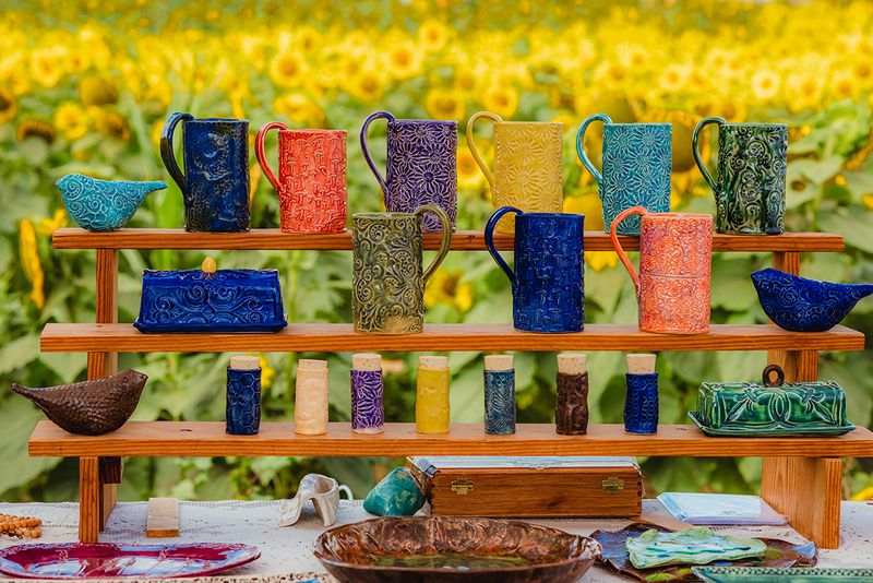 Colorful handmade pottery mugs on a wooden shelf, with the sunflower field behind.